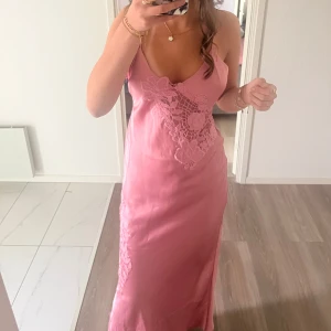 Rosa långklänning med spetsdetaljer - Säljer en elegant rosa långklänning med tunna axelband och vackra spetsdetaljer framtill och på sidan. Klänningen har en feminin siluett och är perfekt för speciella tillfällen.