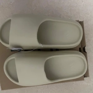 Beige Adidas Yeezy Slide sandaler - Säljer ett par beige Adidas Yeezy Slide sandaler. De har en minimalistisk design med öppen tå och bred rem över foten. Tillverkade i syntetmaterial med räfflad sula för extra grepp. Perfekta för sommaren och riktigt sköna att glida runt i.