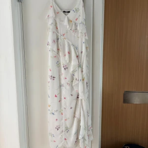 Vit blommig långklänning med volang från Gina Tricot - Säljer en vit långklänning från Gina Tricot med blommigt mönster i pastellfärger. Klänningen har smala axelband, v-ringning och volangdetaljer längs framsidan. Perfekt för sommarens tillfällen! Aldrig använd, storlek 38.
