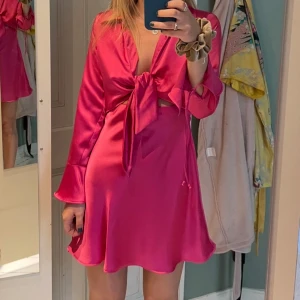 Ceriserosa klänning från Zara - Säljer en ceriserosa  från Zara med knyt framtill. Klänningen har långa ärmar med dekorativa band vid ärmsluten och är tillverkad i ett glansigt, satinliknande material. Perfekt för fest.