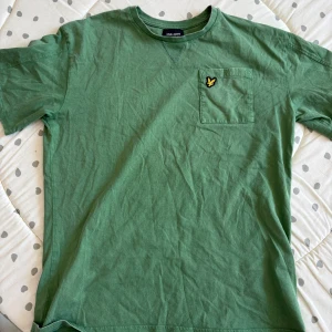Grön t-shirt från Lyle & Scott - Säljer en grön t-shirt från Lyle & Scott Junior i färgen Dark Ivy. T-shirten har en bröstficka med den klassiska gula loggan och är tillverkad i 100% bomull. Perfekt för en avslappnad stil. Storlek (14-15år) väldigt bra skick 