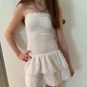 Vit axelbandslös klänning med volang - Från Gina Tricot. Stl XS. Använd någon enstaka gång. Söt vit axelbandslös klänning med smockad överdel och två lager volang med broderade detaljer. Perfekt avslutningsklänning eller för varma dagar och somriga tillfällen. Klänningen har en kort längd och är figurnära upptill. Slutsåld i butik.