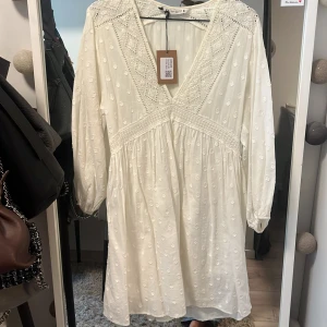Vit klänning med spetsdetaljer från Zara - Superfin vit klänning från Zara med v-ringning och spetsdetaljer. Klänningen har långa ärmar och ett diskret broderat mönster. Perfekt för sommarens tillfällen och har en luftig passform.