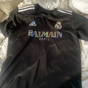 Svart Real Madrid x Balmain fotbollströja från Adidas - Säljer en svart kortärmad fotbollströja med Real Madrid-logga och Balmain Paris-tryck i regnbågsfärger. Adidas-logga på bröstet och klassiska vita ränder på axlarna. Perfekt för dig som gillar unika samarbeten och snygg design pris kan diskuteras .