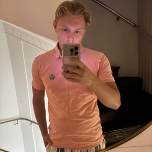 Korallfärgad Selected Homme piké - Snygg korallfärgad pikétröja från Selected Homme med korta ärmar och klassisk krage. Tröjan har en liten broderad logga på bröstet och knappar vid halsen. Perfekt för dig som gillar stilrena och fräscha färger.