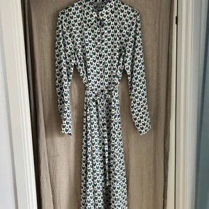 Mönstrad långklänning med knytband - Snygg långklänning med grafiskt mönster i grönt, vitt och rosa. Klänningen har knappar fram, långa ärmar och ett avtagbart knytband i midjan som ger en fin siluett. Perfekt för dig som gillar retrovibbar och unika prints.