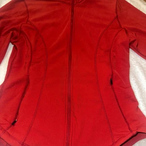 Röd träningsjacka från SOC, stl S - Sportig röd jacka från SOC i storlek S, perfekt för träning. Jackan har hel dragkedja framtill, två sidofickor med dragkedja och är långärmad. Materialet är mjukt och stretchigt syntet, vilket gör den bekväm och följsam. Snygga sömdetaljer ger en modern look.