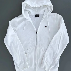 Vit hoodie från Champion med dragkedja - Säljer en clean vit hoodie från Champion i storlek M. Tröjan har huva med snörning, praktiska fickor fram och en diskret Champion-logga på bröstet. Materialet är mjuk bomull med fleece på insidan, perfekt för chill dagar. Dragkedja hela vägen och långa ärmar.