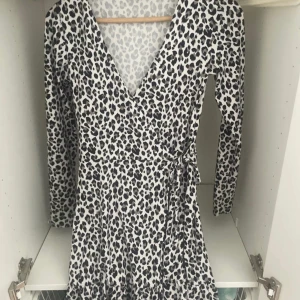 Leopardmönstrad klänning - Snygg kort leopardmönstrad klänning från H&M i storlek XS. Klänningen har omlottmodell med v-ringning, långa ärmar och ett knytband i midjan. Färgen är vit med svarta och grå leopardfläckar. Perfekt för dig som gillar djurmönster och vill sticka ut.