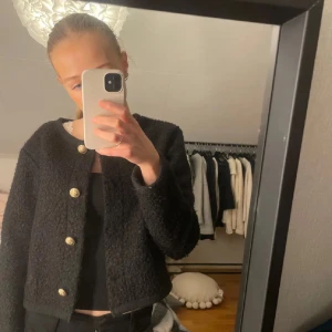 Svart jacka med guldfärgade knappar - Säljer en svart teddyjacka med snygga guldfärgade knappar framtill. Jackan är croppad och har en rak, loose passform med lång ärm. Materialet är fluffigt och mjukt, perfekt för att ge en cool vibe till din outfit.