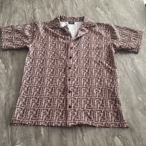Brun mönstrad skjorta från Fendi - Säljer en brun och beige kortärmad skjorta från Fendi med grafiskt rutigt mönster och klassisk krage. Skjortan har knappar framtill och en loose passform som ger en chill vibe. Perfekt för dig som gillar designer och vill sticka ut med coola detaljer.