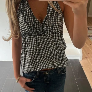 Svartvit rutig ärmlös blus - Supersöt blus i svartvitt rutigt mönster med volangdetaljer längs den djupa v-ringningen. Modellen är ärmlös och har en lätt loose passform som ger en chill vibe. Perfekt till jeans eller kjol för en trendig look. Materialet känns som bomull, skönt och luftigt.