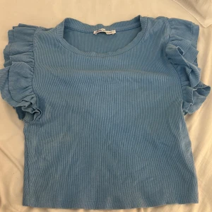 Blå volangtopp från Zara - Supersöt ljusblå topp från Zara i ribbad bomull med korta volangärmar som ger en extra touch. Rund halsringning och croppad passform, perfekt att styla med jeans eller kjol. Säljer då den börjar bli för liten för mig. Storlek S! OBS, katt finns i hemmet!