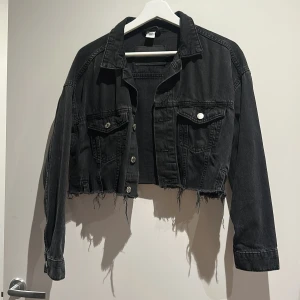 Svart croppad jeansjacka - Säljer en svart croppad jeansjacka med rå kant och slitna detaljer. Jackan har klassisk krage, två bröstfickor med knappar och långa ärmar. Perfekt för dig som gillar en edgy och avslappnad stil.