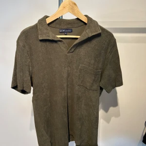 Olivgrön frotté t-shirt från S.C.W. - Olivgrön t-shirt i mjuk frotté från S.C.W. med korta ärmar, v-ringad krage och en bröstficka. Perfekt för chill dagar och har en relaxed vibe med loose passform. Materialet är skönt mot huden och ger en unik look.