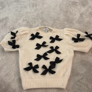 Vit stickad tröja med svarta rosetter - Superfin vit stickad tröja från & Other Stories Paris Atelier i storlek XS. Tröjan har puffiga korta ärmar och är dekorerad med flera svarta rosetter på fram- och ärmsidorna. Mjuk och fluffig känsla, perfekt för dig som gillar unika detaljer och vill sticka ut. 