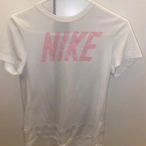 Vit Nike t-shirt med rosa tryck - Snygg vit t-shirt från Nike med stort rosa logotryck på bröstet. Klassisk rund hals och korta ärmar. Perfekt för dig som gillar sportiga och stilrena plagg. Mjuk och skön bomull som passar till många olika outfits.