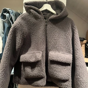 Grå teddyjacka med huva - Mysig brun teddyjacka med stor huva och två rejäla fickor framtill. Jackan har en rak, loose passform och stängs med dragkedja. Perfekt för kalla dagar när du vill vara både chill och varm. 