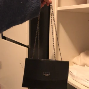 Svart axelväska från PLUS BAG - Snygg svart axelväska från PLUS BAG med stilren design och metallkedja som axelrem.Väskan  är gjord i skinnimitation. 