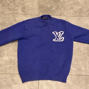 Blå stickad tröja Louis Vuitton - Säljer en blå stickad tröja från Louis Vuitton med stort vitt LV-logo på både fram- och baksidan. Tröjan har rund hals, ribbade muddar och lång ärm. Perfekt för dig som vill ha en lyxig och snygg statement piece i garderoben.