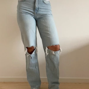 Ljusblå ripped jeans med raka ben - Säljer ett par ljusblå jeans med raka ben och snygga slitningar vid knäna. Jeansen har hög midja och klassisk femficksdesign. Perfekta för dig som gillar en avslappnad och trendig stil.