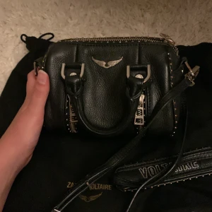 Svart handväska Zadig & Voltaire skinn - Cool svart handväska från Zadig & Voltaire i äkta skinn med silverfärgade detaljer och nitar längs kanterna. Väskan har två handtag, avtagbar axelrem och snygga dragkedjor framtill. Perfekt storlek för det viktigaste och har en edgy vibe med metallvinge på framsidan. väskan är lite rostig vid dragkedjan.❤️