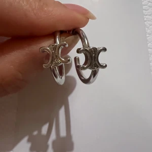 Celine Triomphe örhängen i silver - Säljer ett par chunky hoop-örhängen från Celine med den ikoniska Triomphe-loggan framtill. Örhängena är i blankt silverfärgat metall och har en rund, tjock form som ger en cool vibe. Perfekta för dig som gillar statement-smycken och designeraccessoarer.