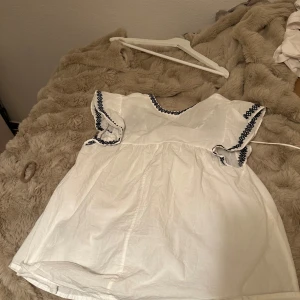 Vit kortklänning med broderi och snörning - Supersöt vit blus med svarta broderade detaljer och snörning vid halsen. Blusen har korta ärmar med volang. Tyvärr för stor för mig. Kan diskutera pris💘💘