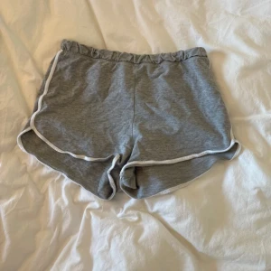 Grå mjuka shorts med vit kant - Superbekväma grå shorts med elastisk midja och vit kant längs sidorna. De har en loose passform och är gjorda i mjuk bomull, perfekta för chill dagar eller träning. Snygg sportig look med rundade benkanter.