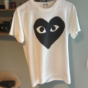 Vit Comme des Garçons Play t-shirt - Säljer en vit t-shirt från Comme des Garçons Play i storlek M. Framsidan har det ikoniska svarta hjärtmotivet med ögon. T-shirten är i mjuk bomull och har klassisk rund hals och korta ärmar. Perfekt för dig som gillar streetwear och coola prints.