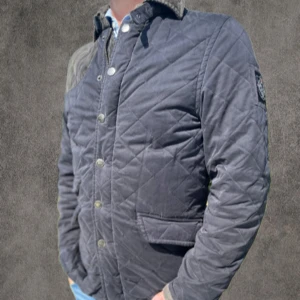 Quiltad fieldjacka från Morris, mörkgrå S - Snygg quiltad fieldjacka från Morris i mörkgrått med klassiskt emblem på ärmen. Jackan har knappar framtill, två fickor och krage i manchester. Materialmix av bomull och polyester, perfekt för höst och vår. Stilren och tidlös design som passar till det mesta.