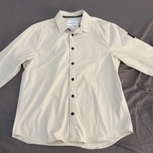 Beige overshirt från Calvin Klein Jeans - Snygg beige overshirt från Calvin Klein Jeans i bomull med svart CK-logga på ärmen. Rak passform med svarta knappar framtill och klassisk krage. Perfekt för dig som gillar stilrena och enkla plagg med en modern touch.