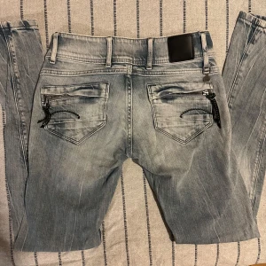 G-Star ljusgråblå jeans med låg midja – strl 24/30 - G-Star ljusgråblå jeans med låg midja – strl 24/30 Ljusgråblå jeans från G-Star med låg midja, dragkedjor på bakfickorna och läderpatch i midjan. Slim passform och klassisk femficksdesign. Materialet är slitstarkt jeans med modern, lätt tvättad look, perfekt för streetwear. Skick: Nästan nya, använda få gånger