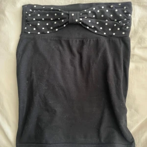 Svart topp med prickig rosett - Supersöt svart topp med en stor rosett framtill i svart tyg med vita prickar. Toppen är figurnära och har en enkel, stilren design som får extra attityd av den prickiga detaljen. Perfekt för dig som gillar att sticka ut med detaljer.