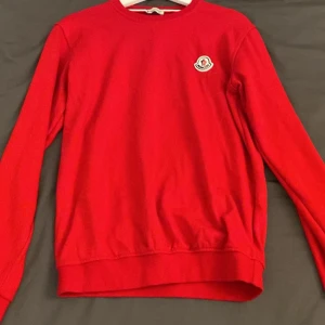 Röd långärmad tröja från Moncler - Snygg röd långärmad tröja från Moncler med rund hals och klassisk logga på bröstet. Tröjan har en enkel och clean design med ribbade muddar vid ärmslut och nederkant. Perfekt för dig som gillar stilrena märkesplagg.