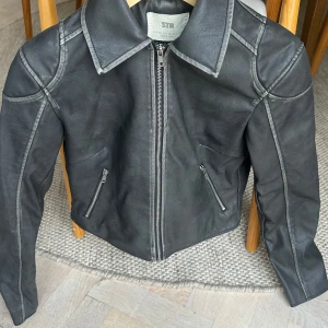 Svart skinnjacka från STR - Snygg svart skinnjacka från STR med markerade sömmar och klassisk krage. Jackan har två dragkedjeförsedda fickor framtill och hel dragkedja. Stilren design med långärmade ärmar och en något kortare passform som ger en cool vibe.