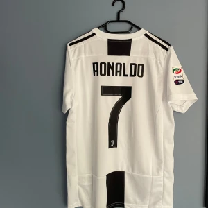 Juventus Ronaldo 7 matchtröja Adidas - Juventus fotbollströja med Ronaldo 7 på ryggen, Tröjan har klassiska svarta och vita ränder, korta ärmar och Serie A-märke på ena ärmen. Materialet är lätt och andas, perfekt för match eller träning. Jeep-logga på bröstet och tre stjärnor ovanför klubbmärket.
