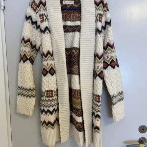 Mönstrad stickad kofta med bälte - Säljer en mysig och lång stickad kofta från Odd Molly med bälte i midjan. Koftan är vit med mönster i rött, svart, beige och grönt, har blommiga och zigzag-detaljer samt grov ribbad krage. Perfekt för dig som gillar boho-vibe och vill ha något extra till hösten och vintern.