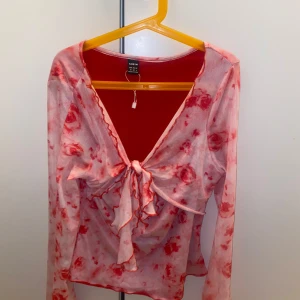 Blommig meshblus med knyt från SHEIN - Säljer en transparent meshblus från SHEIN i storlek M. Blusen har ett blommigt mönster i rosa och rött, v-ringad hals och snygg knytning framtill. Långärmad och med volangdetaljer längs kanterna för extra vibe. Perfekt för dig som gillar att sticka ut!
