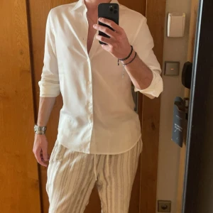 Vit skjorta från J.Lindeberg - Snygg vit skjorta från J.Lindeberg med klassisk krage och diskret broderad logga vid nederkanten. Skjortan har lång ärm och knappar framtill. Tillverkad i mjuk bomull som känns skön mot huden. Perfekt för dig som gillar stilrena och cleana plagg.