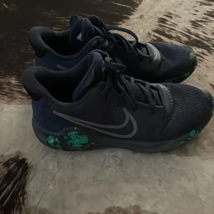 Mörkblå Nike sneakers med gröna detaljer - Säljer ett par mörkblå KD trey emerald 5 sneakers med snygga gröna mönster på sulan och silverfärgad swoosh på sidan. Skorna har snörning och rund tå, tillverkade i syntetmaterial med mesh för extra komfort. Perfekta för dig som gillar sportig stil och coola färgkombinationer.