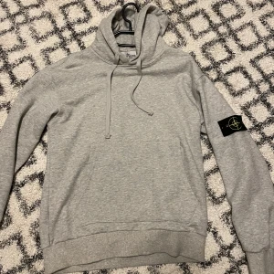 Grå hoodie från Stone Island - Säljer en grå hoodie från Stone Island med klassisk patch på vänster ärm. Tröjan har huva med snörning, känguruficka och ribbade muddar. Perfekt för dig som gillar streetwear och vill ha en clean look med snygg branding.