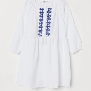 Vit kortklänning med blå broderi H&M - Supersöt vit kortklänning från H&M i storlek 134, med lång ärm och blå broderade detaljer framtill. Klänningen har knappar vid halsen och är gjord i mjuk bomull med diskret struktur.