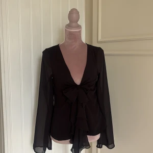 Svart cover-up från Nakd - Svart cover-up från NA-KD med djup v-ringning och knytning framtill. Långa, transparenta ärmar och volangdetaljer. Blusen är gjord i lätt polyester som faller snyggt och känns luftig. Perfekt för dig som gillar statement-plagg.