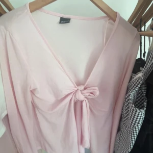 Ljusrosa tröja från Gina Tricot - Supersöt ljusrosa topp från Gina Tricot i mjuk bomull. Toppen har lång ärm, v-ringning och en snygg knytning framtill som ger en chill vibe. Perfekt att styla med jeans eller kjol för en trendig look.