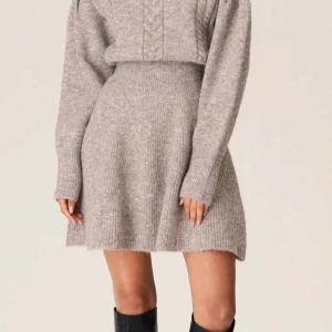 Beige stickad klänning från Adoore - Supermysig beige stickad klänning från Adoore med kabelstickade detaljer på överdelen och ribbad midja. Klänningen har lång ärm, rund hals och är kort i modellen. Perfekt för dig som gillar en chill och snygg look under höst och vinter.