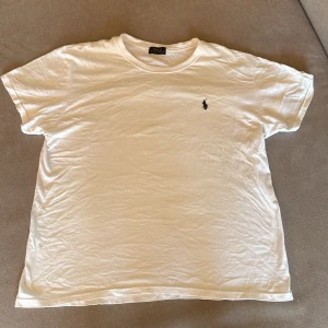Vit t-shirt från Polo Ralph Lauren - Vit t-shirt från Polo Ralph Lauren med klassisk svart broderad logga på bröstet. Modellen är normal passform med rund hals och korta ärmar. Mjuk och skön bomull som känns chill och enkel att matcha med jeans eller shorts.