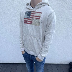 Ljusgrå hoodie med amerikansk flagga - Säljer en ljusgrå hoodie med tryck av amerikansk flagga på bröstet. Tröjan har huva, känguruficka och långa ärmar. Perfekt för chill dagar och enkel att matcha med jeans. Materialet känns mjukt och bekvämt mot huden.
