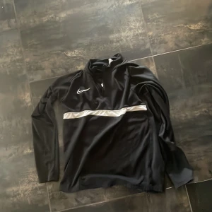Svart Nike half zip tröja - Svart långärmad tröja från Nike med vit logga på bröstet och en silverfärgad rand över magen. Tröjan har half zip och är gjord i mjukt polyester-material, perfekt för träning eller chill. Snygg och sportig look!