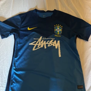 Blå Brasil Nike x Stüssy fotbollströja M - Snygg blå Brasil fotbollströja från Nike i samarbete med Stüssy. Tröjan har korta ärmar, Brasil-logga med stjärnor, gul Nike swoosh och stor vit Stüssy-logga på bröstet. Tillverkad i Dri-Fit material som andas och håller dig torr. Perfekt för dig som gillar streetwear och fotboll. Fantastiskt skick 10/10 